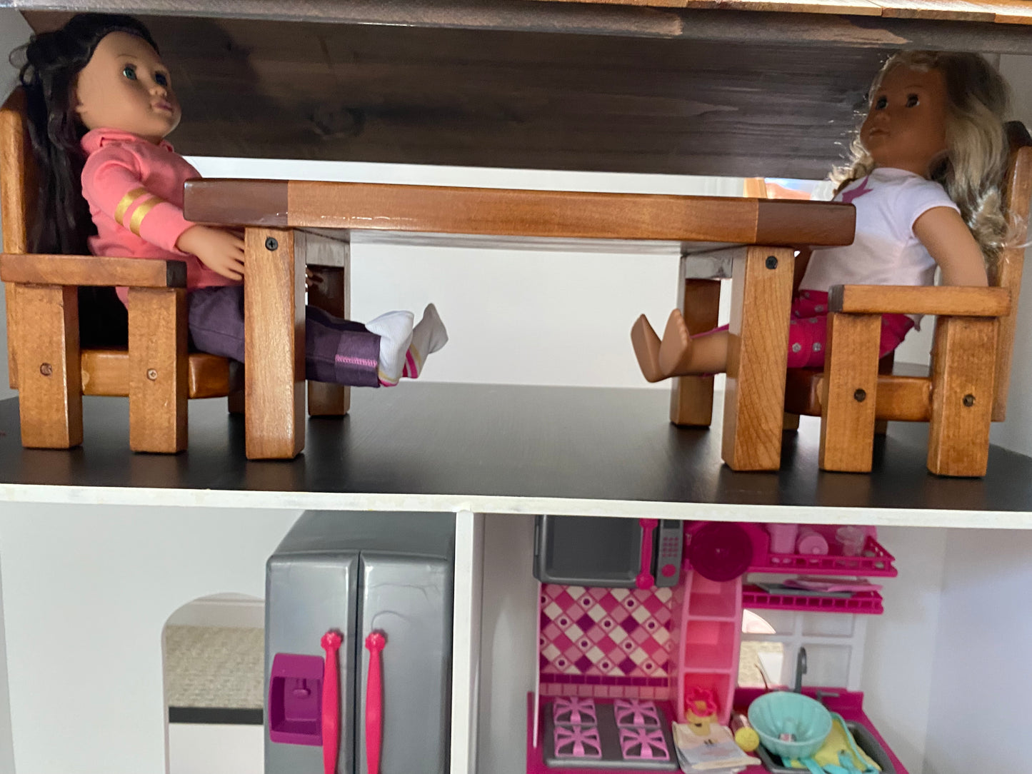 Two dolls sitting at a small wooden table with a toy kitchen set in the background.